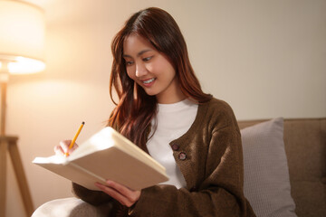 Young asian women sitting on comfortable the couch to reading literature book and writing diary...