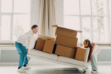 Happy young family couple man and woman lift and carry sofa with cardboard boxes in new home....