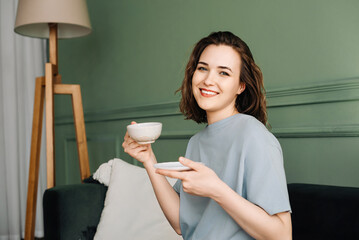 A cheerful pretty young woman with a cup of hot drink sitting on a couch enjoying a vacation. Couch...