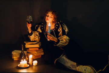 Young adult woman dressed in a medieval dress sitting near ancient books and candle light with bottle of red wine and holding a glass of wine in dark