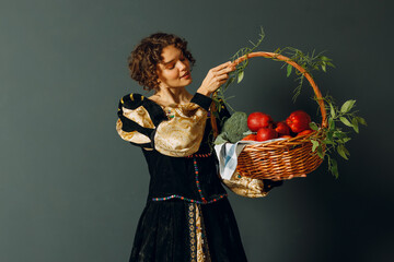 Portrait of a young adult woman dressed in a medieval dress holding a basket with vegetables and fruits in her hands