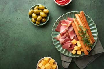 Slices of prosciutto or jamon. Delicious grissini sticks with prosciutto, cheese, rosemary, olives on green plate on dark background. Appetizers table with italian antipasto snacks Top view copy space