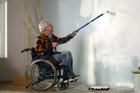 The Picture Shows The Room In Which The Repair Is Carried Out. An Elderly Man In A Wheelchair. He Is A Repairman Who Paints A Roller On A Long Stick Of Paint On The Wall