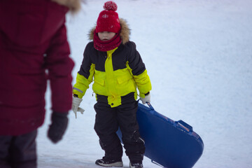 A little boy in a warm yellow jacket carries a sled up the hill to slide down