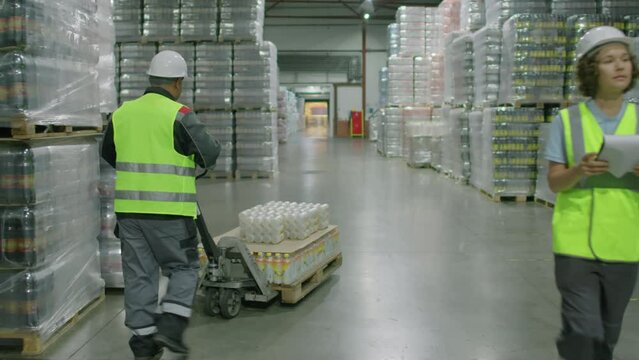 Tracking shot of male storage operator pulling cart with pallet of beverage bottles while preparing freight for shipping