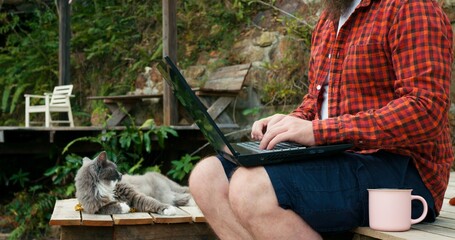 Freelancer blogs on laptop or does work with stunning view of jungle. Cat rests on wooden pier next to man who drinks tea from cup in jungle. Remote work concept in jungle by river.