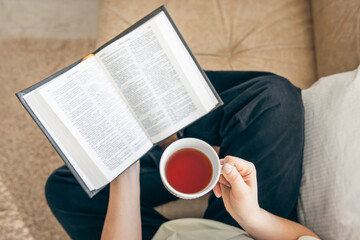 Fototapeta na wymiar Close up, young woman drinking hot tea and reading favorite paper book, top view.