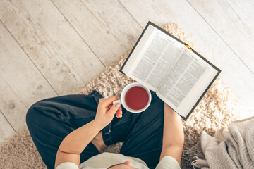 Top view, woman holding mug of tea while reading a book on the floor.