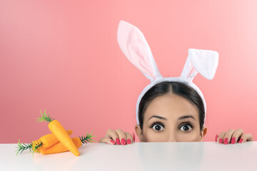 Beautiful young woman with pink bunny ears and toy carrot on pink background. Minimal Easter concept.