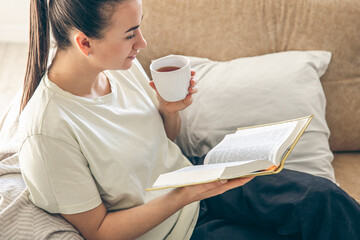 Young woman drinking hot tea, reading favorite paper book at home.