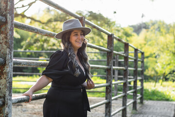 beautiful latina girl posing next to an iron fence, smiling and looking very happy, with a hat and dress.