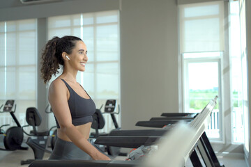 Fototapeta na wymiar Happy healthy hispanic woman practicing a cardio exercise by running and walking on treadmill machine in fitness and using fitness tracker or smartwatch to track workout progress.