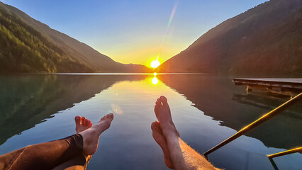 Couple enjoying allure of sunset at alpine lake Weissensee in remote Austrian Alps in Carinthia. Hanging legs of people. Embrace serene ambiance as tranquil surface of water amplifies sun gentle rays