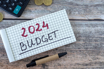 2024 budget, handwritten text in notebook with coins, calculator, and pen on wooden table. Top...