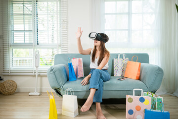 Asian young woman with shopping bags around wearing modern virtual reality headsets having experience in shopping online in living room at home, Happy female with VR headset on sofa