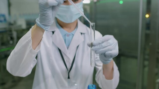 Tilt up shot of woman in white coat and protective gloves testing water in PET bottle at beverage factory before delivery