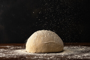 Pastry chef sprinkles flour on fresh raw dough for bread or pizza on a dark background.