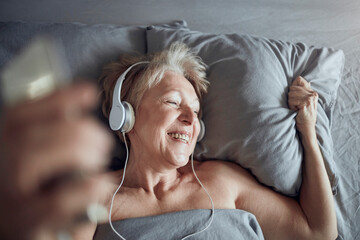 Happy senior woman listening and enjoying relaxing music in bed