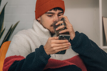 young man at home warmly drinking looking at mobile phone