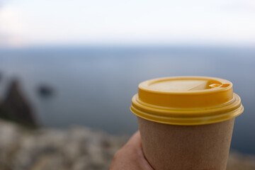 Hand holding Yellow cup with lid, coffee against a backdrop of a blue sky and sea. Illustrating cup and beverage