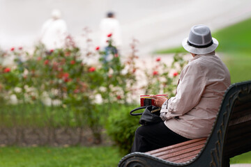 An elderly woman in a hat in the city square on a bench. She is holding a half-closed book. Copy...