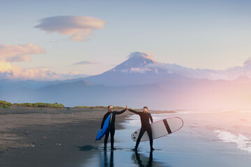 Group of friends means in wetsuit going to surf at beach. Lifestyle Extreme Surfer winter surfing in North ocean Kamchatka Russia. Friendship adventure travel sport concept - obrazy, fototapety, plakaty