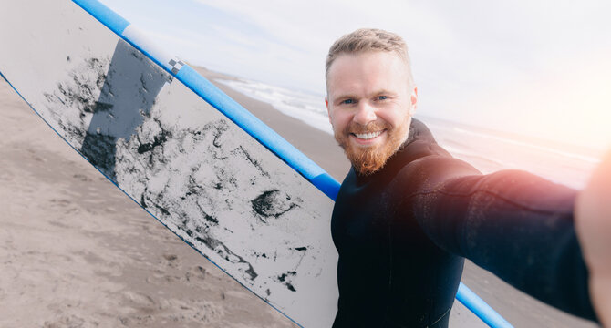Extreme selfie photo Surfer man in wetsuit with surfboard, winter surfing in ocean, sunlight. Adventure travel sport concept