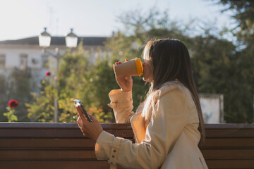 Woman drinks from cup on wooden bench. She is wearing a white shirt enjoying her beverage. The bench is located in a park setting, with trees in the background.