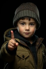 Portrait of a blue-eyed little boy in winter hat, looking and pointing at camera with his index finger on black background.