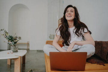Happy woman working on laptop while sitting on a mustard yellow couch in a room with minimalist...