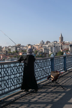 An Arab woman in a black abaya stands and fishes on the Galata Bridge in Istanbul. The urban development of Beyoglu district is in the background.