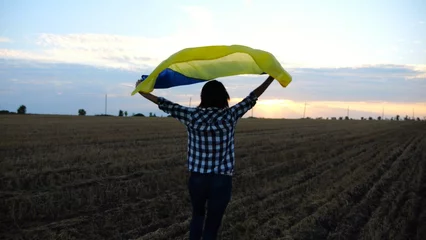 Foto op Canvas Ukrainian woman running with raised flag Ukraine above her head on wheat field at sunset. Lady jogging with national blue-yellow banner on barley meadow at sunrise. Victory against russian aggression. © ExpressVectors