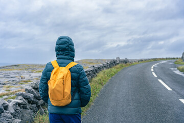 Unknown female hiker with hood on and yellow backpack on her back walks on a lonely road in the...