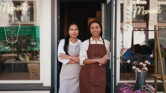 Smiling multicultural waitresses in aprons welcoming guests at door of cafe 
