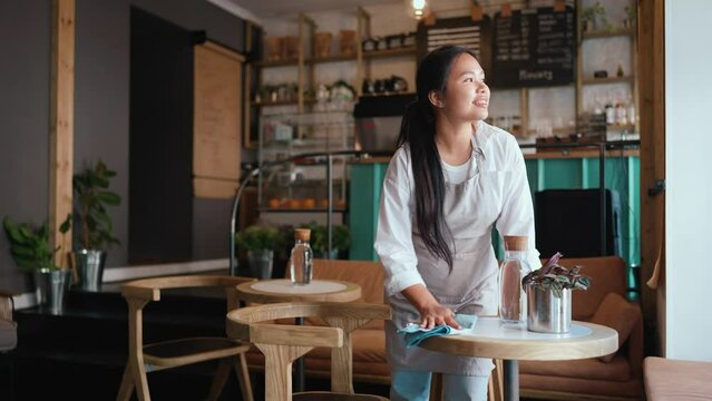 Smiling asian woman waitress in apron wiping tables in cafe
