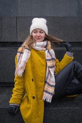 A stylish elegant woman in a yellow coat and in a white hat sits on a dark granite monument....