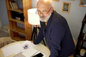Suspicious concerned mature trader standing in stylish cardigan in cabinet putting hands on table with smartphone, looking up at camera, inventing plan for investment, analyzing stock exchange market