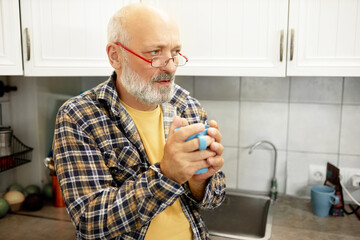 Side view of cold senior man in plaid shirt and glasses warming his hands holding hot drink in blue...