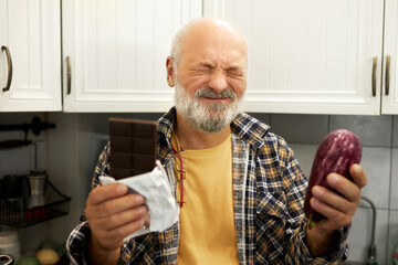 Funny indoor portrait of man with closed eyes holding chocolate bar and aubergine, trying to chose...