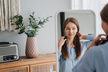 Confident woman in blue shirt adjusting her earring in front of a mirror, with a vintage radio and houseplant.