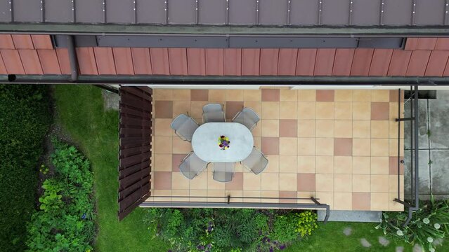 terrace by the house with a beautiful arranged flower in a vase. paving tiles brown and yellow. oval table, metal gray chairs. metal railing drone view. a flower bed surrounds the lawn