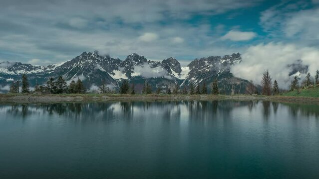 Timelapse of the moving clouds in the mountains of Wilder Kaiser with snow in Austria Tyrol, mirroring on the surface of the lake of Astberg during spring with clouds in the blue sky
