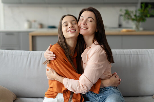 Mother and teenage daughter embracing, enjoying moment together, sitting with closed eyes on couch at home, trusted relationships between mom and teen girl