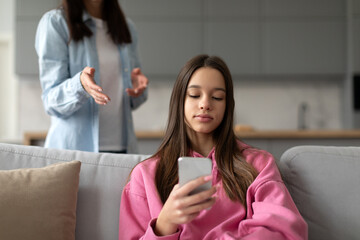 Angry mother having trouble with teenager girl, sitting on sofa at home and playing video games on...