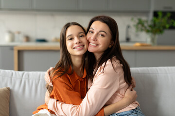 Loving mother hugging embracing her happy teen daughter at home, expressing love to teenage child, sitting together on couch and cuddling