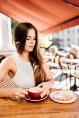 Portrait of happy woman sitting in a cafe outdoor drinking coffee. Woman while relaxing in cafe at table on street, dressed in a white T-shirt and jeans