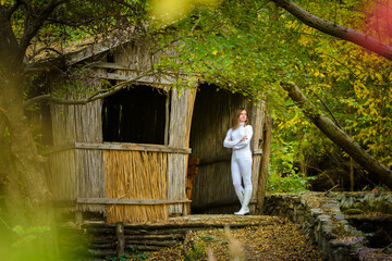 A young beautiful girl in white clothes stands near an old abandoned house in the forest