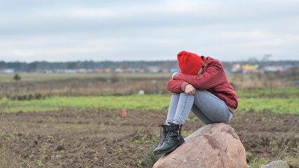 A sad teenage girl sits on a rock in a field and cries.