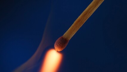 Macro shot of a burning match against a blue studio background. The flame of the burning match illuminates the dark space. The burning match is enveloped in an orange flame.