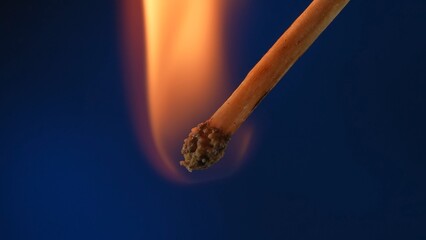 Macro shot of a burning match against a blue studio background. The flame of the burning match illuminates the dark space. The burning match is enveloped in an orange flame.